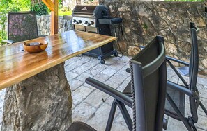 Un muro de piedra, una parrilla, una mesa de madera y sillas de metal.