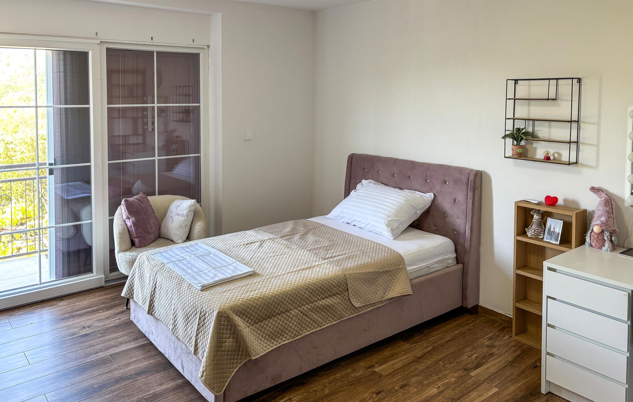 A bedroom with a bed, a chair, a desk, a shelf with decorative items, and a sliding glass door.