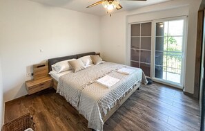 Un dormitorio con cama, mesita de noche, ventilador de techo y un ventanal grande.