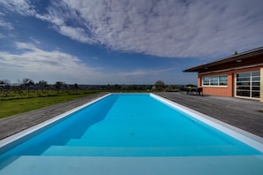 Una piscina rectangular con muelle de madera alrededor, un edificio con amplias ventanas a la derecha, y un cielo despejado con nubes.