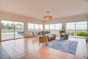 Un salón amplio con un sofá gris, dos sillones, una mesa de centro y una alfombra azul. Grandes ventanales ofrecen vistas al exterior.