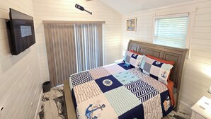 Un dormitorio con ventilador de techo de madera, cama con ropa de cama de temática náutica, televisor montado y ventana con persianas.