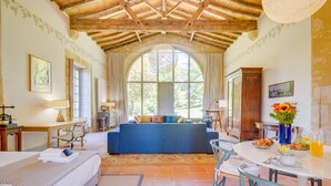 Quarto com cama, sofá, mesa de jantar com cadeiras, uma grande janela em arco e teto de madeira.