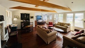 Un amplio salón con vigas de madera, chimenea, zona de comedor y ventanas grandes.