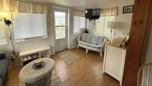 Ein gemütliches Zimmer mit einer Couch, einem kleinen Tisch, einem an der Wand befestigten Fernseher und einem Fenster mit Jalousien.
