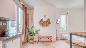 A compact room with a bed, a small table, a bench, a refrigerator, a potted plant, and a wall-mounted decorative item.