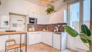 A modern kitchen with white cabinets, a wooden countertop, and a small refrigerator. There is a bar stool and a large plant.