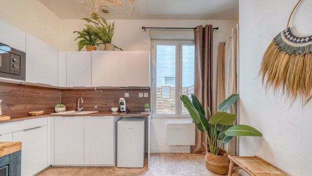 A modern kitchen with white cabinets, a built-in microwave, and a small refrigerator. There is a window with curtains, a potted plant, and a bench.
