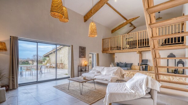Amplia sala de estar con un gran ventanal corredero que da a un patio exterior, un entrepiso con escalera de madera y una zona de estar acogedora con sofá y sillones.