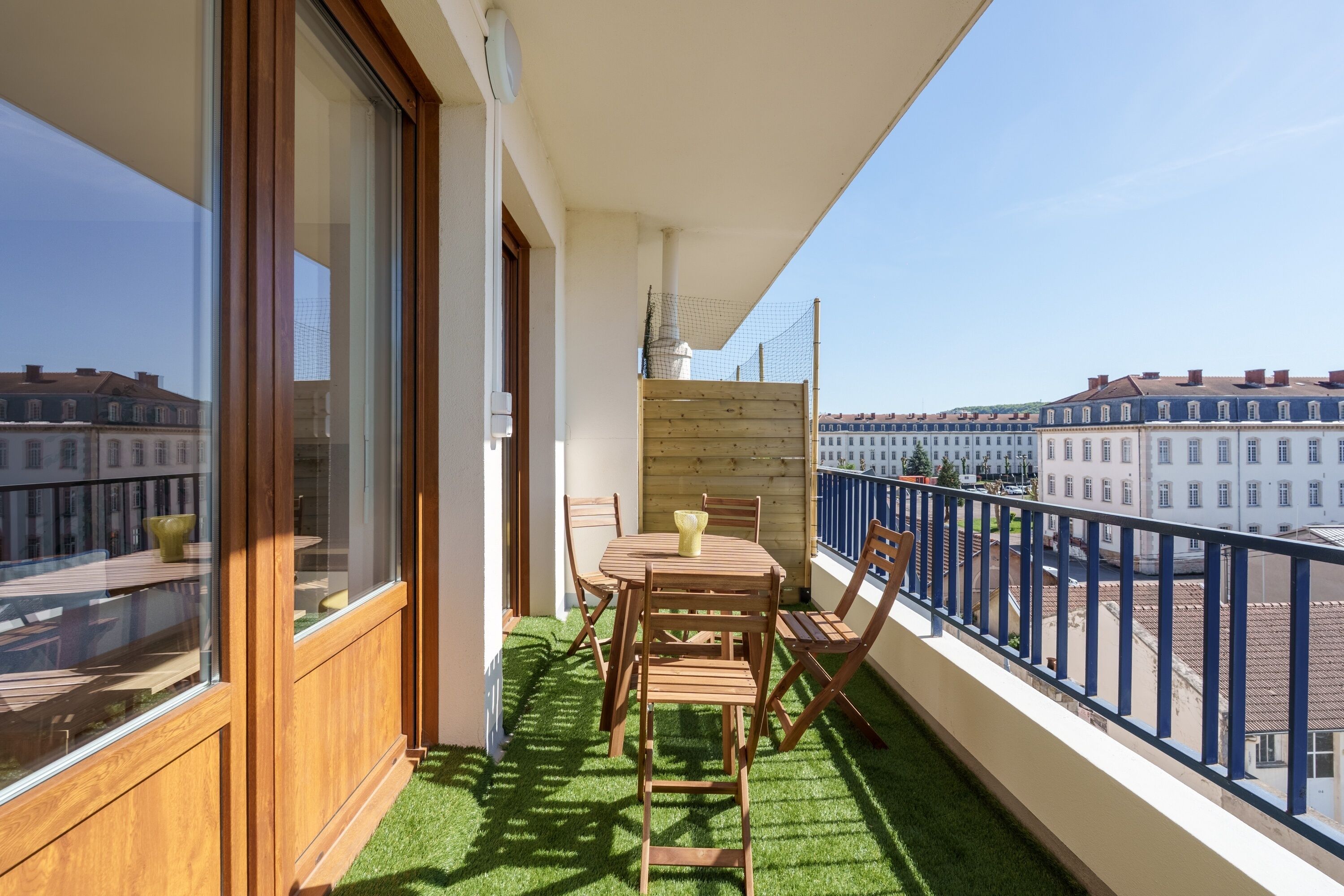 A balcony with a wooden table and chairs, a small wooden fence, and a view of buildings.