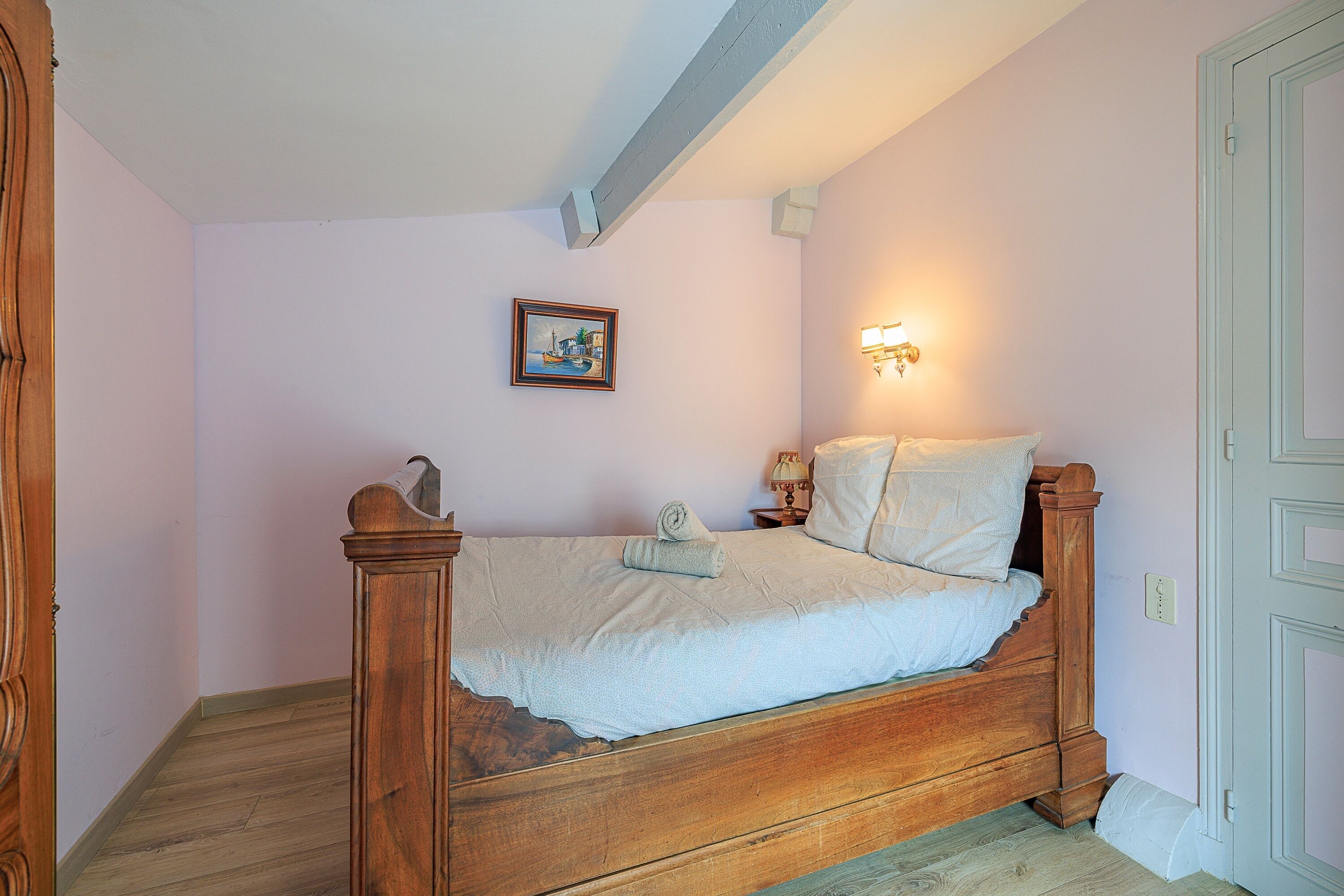 A bedroom with a wooden bed, a small bedside table, a wall-mounted light, and a framed picture on the wall.