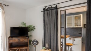 Ein Wohnzimmer mit Fernseher, einem Ventilator, einem kleinen Tisch mit Lampe und einer Küchenzeile mit Barhocker.