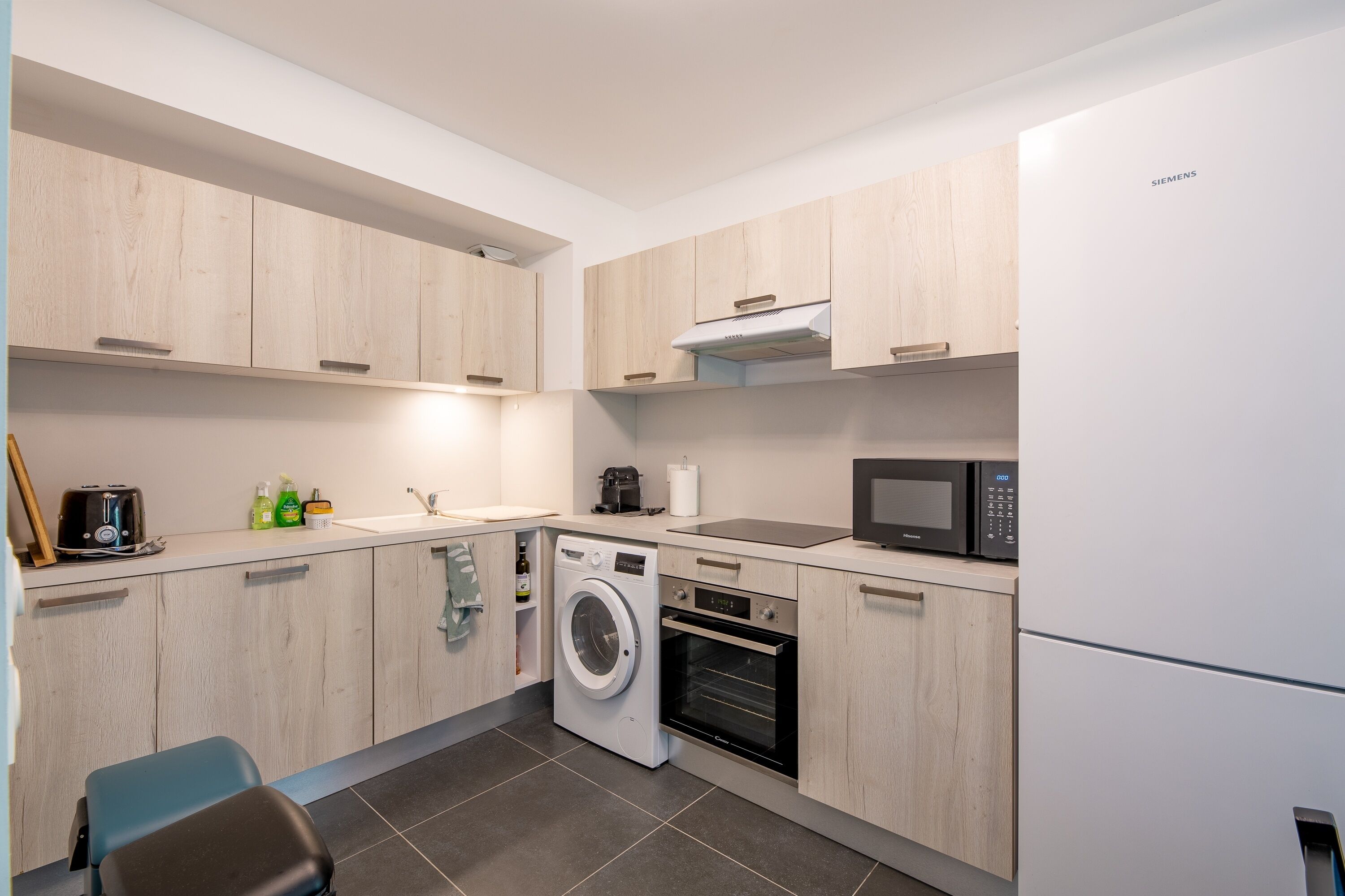 A modern kitchen with wooden cabinets, a washing machine, an oven, a microwave, and a refrigerator.