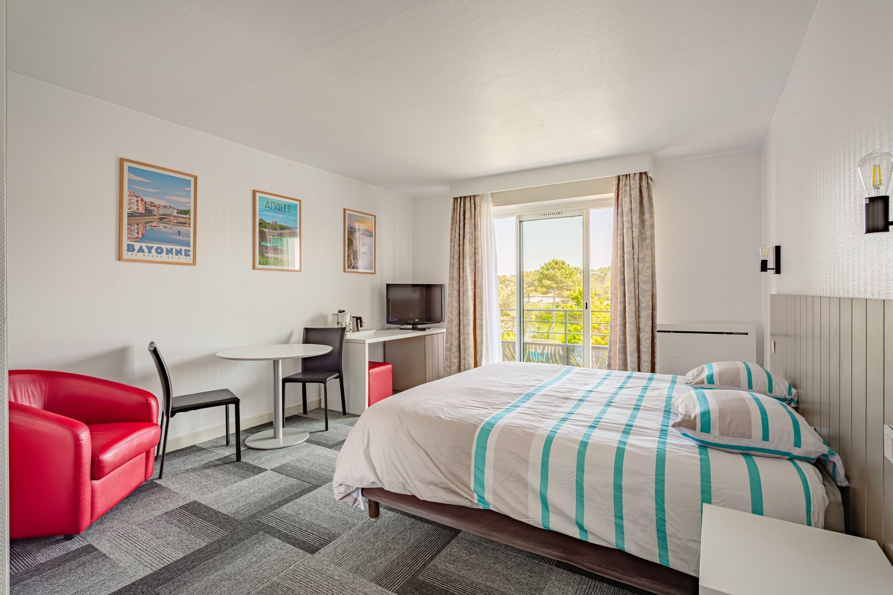A hotel room with a bed, a desk, a TV, a red chair, and a balcony with a view.