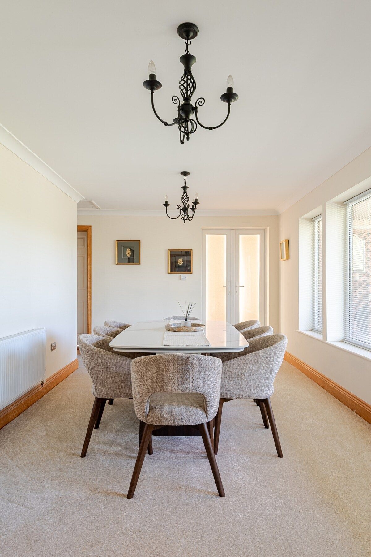Comedor con una mesa blanca y cuatro sillas tapizadas, una lámpara de araña negra y cuadros en la pared.