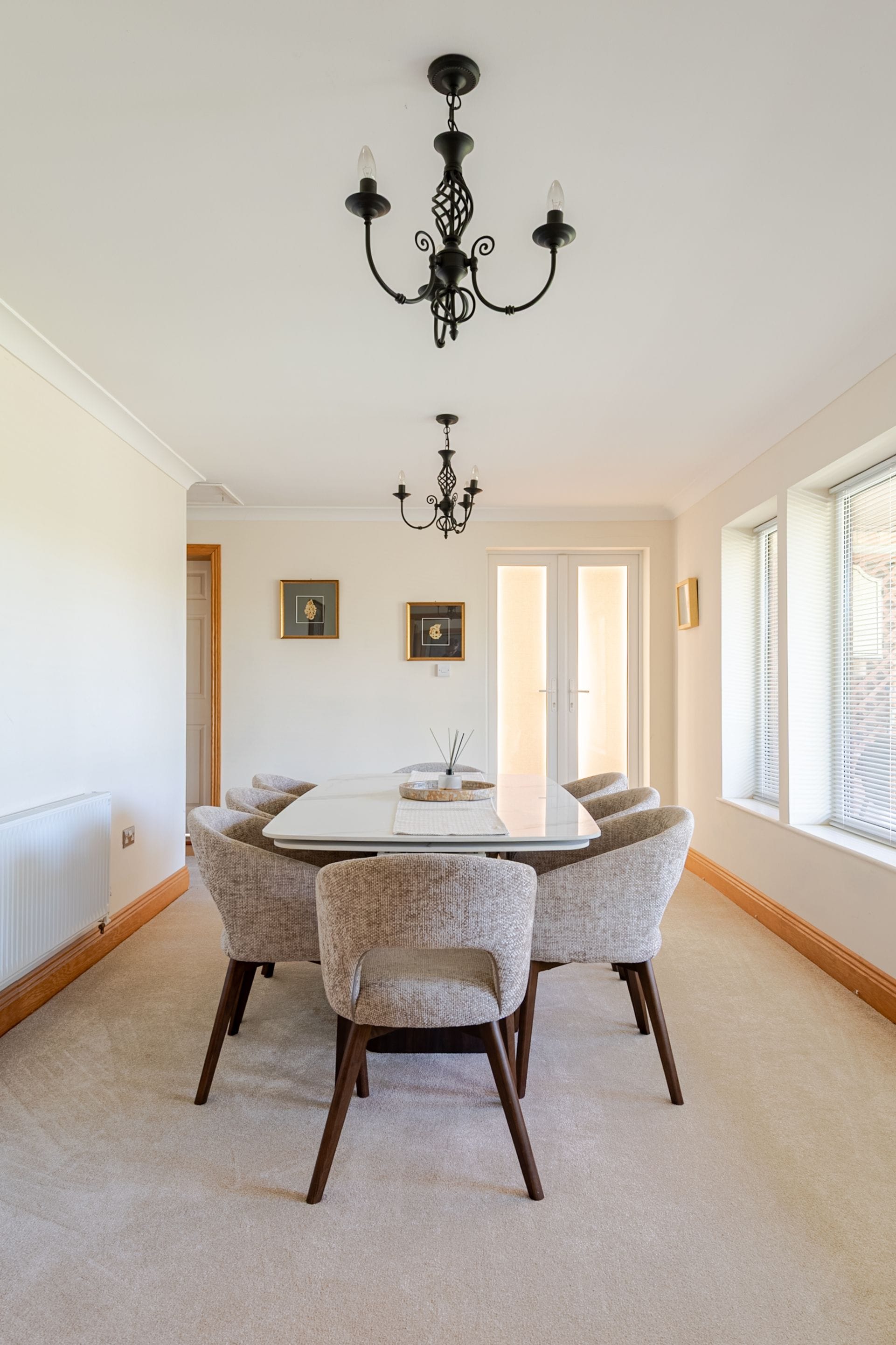 Une salle à manger avec une table blanche, quatre chaises rembourrées, un lustre noir et des tableaux encadrés accrochés aux murs.
