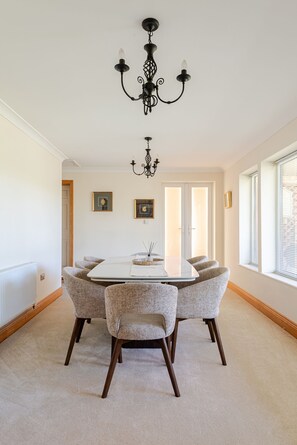 Un comedor con una mesa blanca, cuatro sillas tapizadas, un candelabro negro y una ventana con persianas.