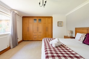 Un dormitorio con un armario grande, una cama con colcha a cuadros, una mesita de noche con lámpara y una ventana con cortinas.