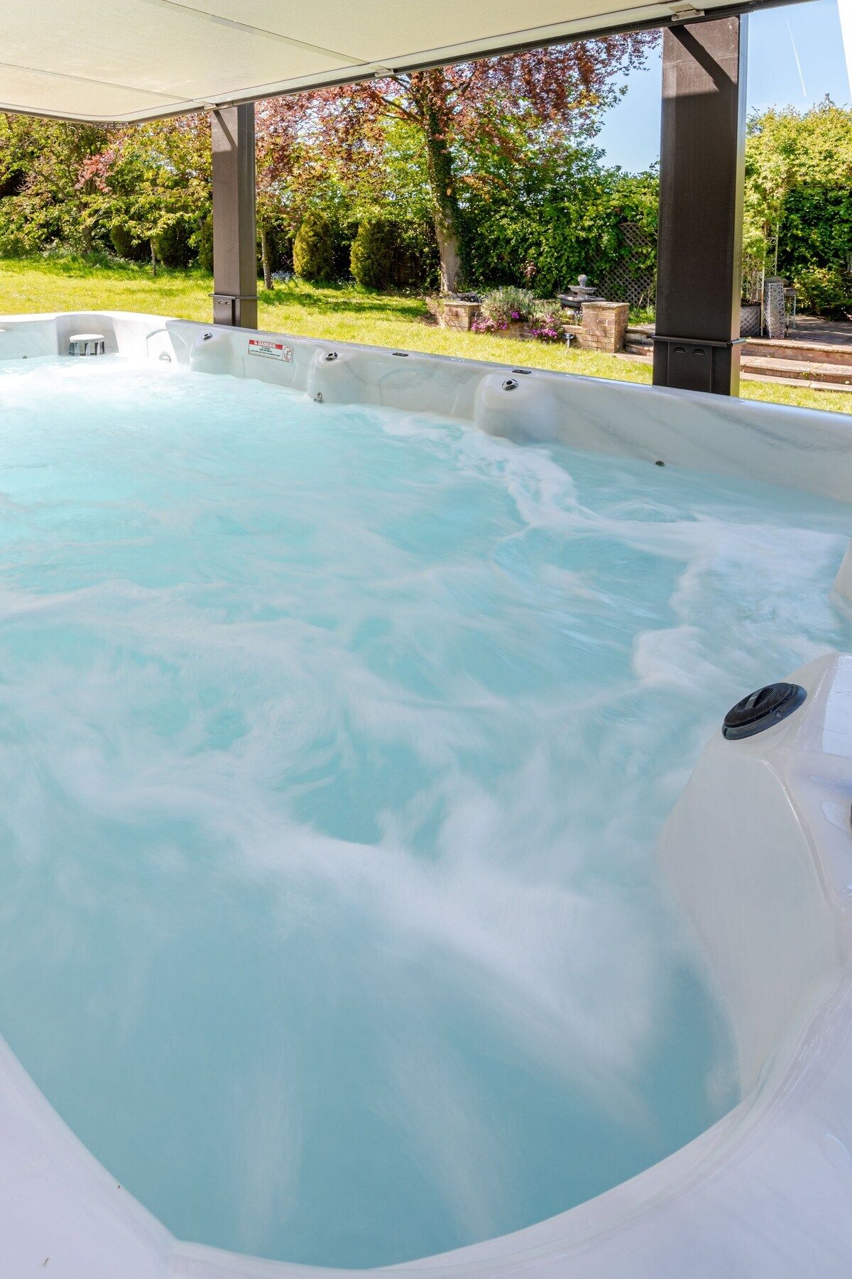 Een hottub onder een overdekte ruimte, met een gazon en bomen op de achtergrond.