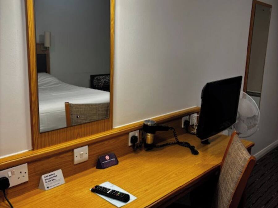 A hotel room with a wooden desk, a computer, a chair, a bed, a mirror, and a bedside table with a lamp.
