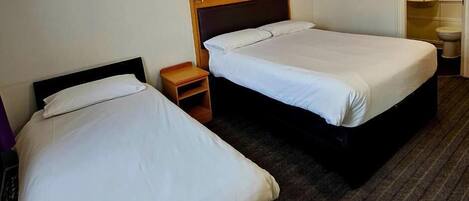 Two single beds in a hotel room with a headboard, a small wooden nightstand, and a bathroom visible through an open door.