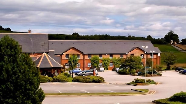 A hotel with a parking lot, a covered outdoor area, and a hilly landscape in the background. - Monifieth Farm Hotel (Dundee)