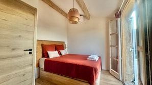 A bedroom with a wooden bed, a red bedspread, a hanging light fixture, and a large wooden door. - House near downtown Saint-Tropez (Gassin)