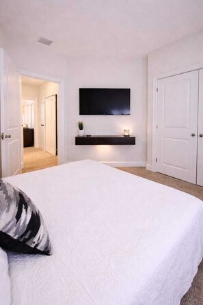 Un dormitorio con una cama grande, un televisor montado en la pared, una mesita de noche con una planta y una puerta que da a otra habitación.