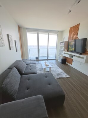 Un salon moderne avec un grand canapé d’angle gris, un téléviseur à écran plat posé sur un meuble de télévision blanc, et une vue sur la mer grâce à de grandes portes coulissantes.