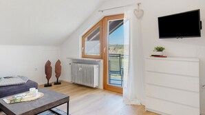 A modern room with a sloped ceiling, a large window, a flat-screen TV, a radiator, and a sliding glass door leading to an outdoor area. - Dog-Friendly Holiday Apartment with 2 Balconies and Wi-Fi (Bräunlingen)