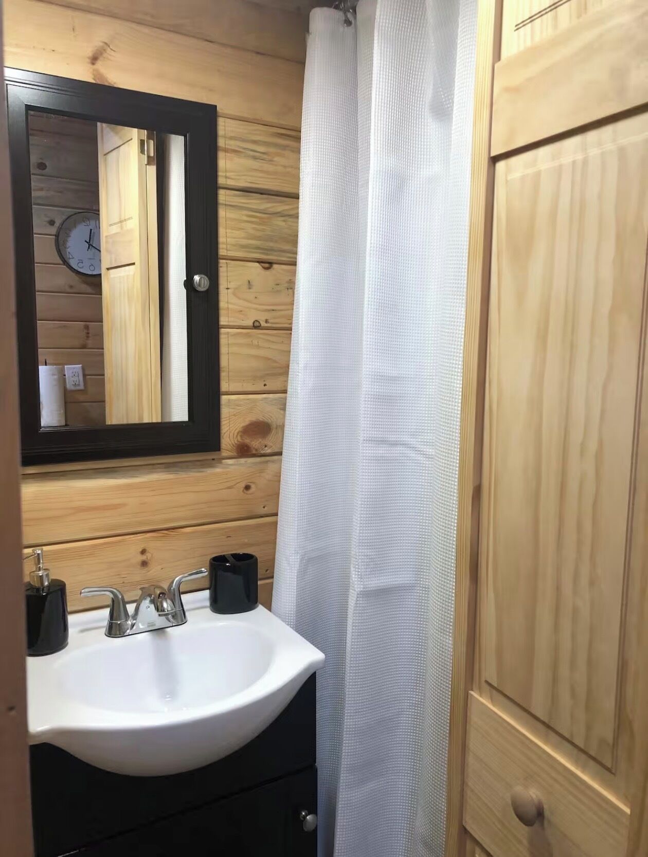 A bathroom with a wooden wall, a white sink, a mirror, a clock, and a white shower curtain.