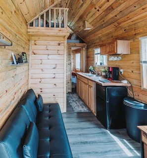 Interior de una cabaña con sofá, zona de cocina y escalera que sube a un altillo.