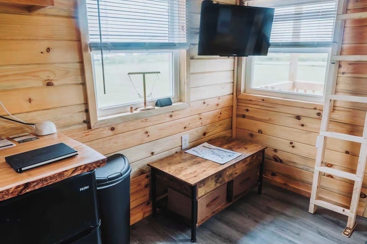 Una cabaña con paredes de madera, un escritorio con una computadora portátil, un televisor y una escalera.
