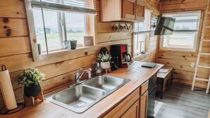 A wooden cabin kitchen with a double sink, wooden cabinets, a coffee maker, and a window overlooking a grassy area. - Cozy tiny home with Sunrise Views in the Heart of Finger Lakes (Penn Yan)
