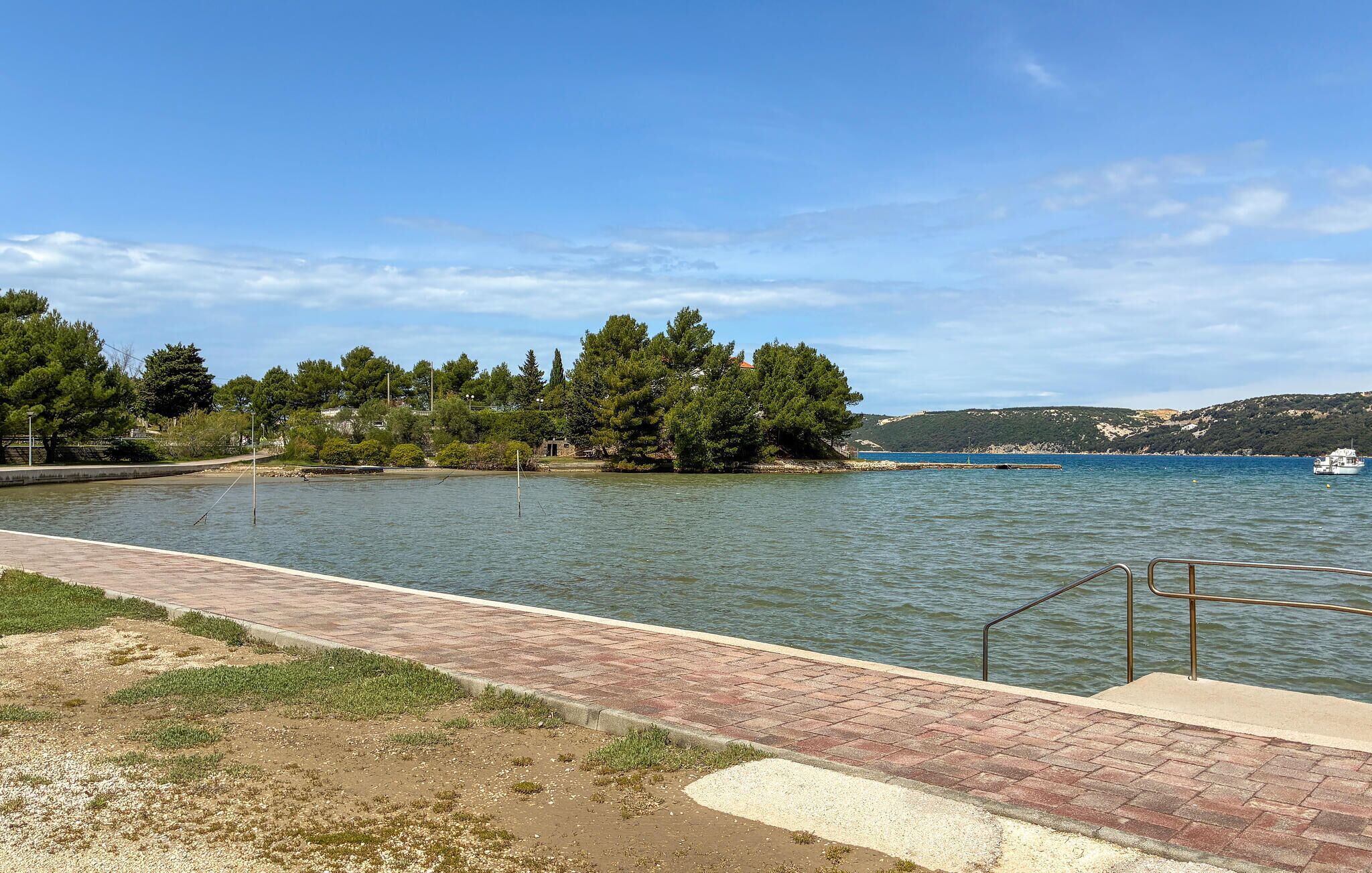 Une promenade en bord de mer, avec un chemin pavé de briques, des arbres et un bateau visible au loin.