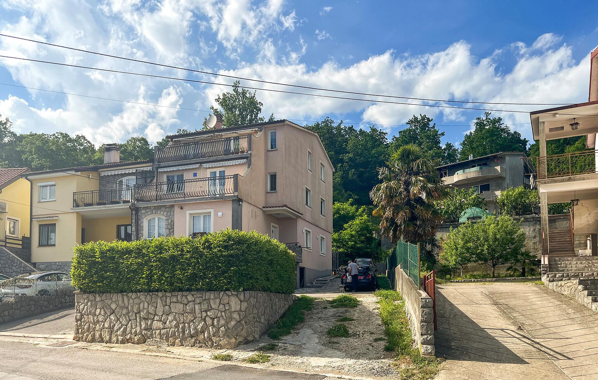 A residential street with multi-story buildings, a stone wall with greenery, and a clear sky.