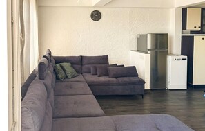 A living room with a sectional sofa, a wall clock, and a kitchen area with a refrigerator. - 2 bedroom awesome apartment in Kastav (Kastav)