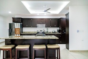 A modern kitchen with dark cabinets, a central island with stools, a ceiling fan, and built-in appliances. - Casa Descanso (Puerto Peñasco)