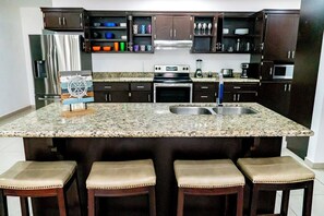 A kitchen with dark cabinets, a stainless steel refrigerator, an oven, a sink, and a coffee maker. There are four stools with cushioned seats. - Casa Descanso (Puerto Peñasco)