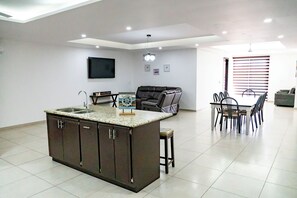 A modern kitchen with a central island, dining area with chairs, and a flat-screen TV mounted on the wall. - Casa Descanso (Puerto Peñasco)