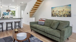 A modern living space with a staircase, a green sofa, a cityscape painting, and a kitchen area with stools. - Beautiful 2-bedroom townhome downtown Raleigh  Walk to Everything (Raleigh)