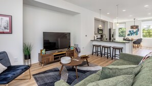 A modern living room with a flat-screen TV, a sofa, a coffee table, and a kitchen area with a dining table and bar stools. - Beautiful 2-bedroom townhome downtown Raleigh  Walk to Everything (Raleigh)