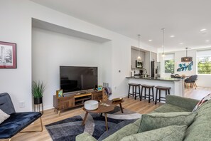 Ein modernes Wohnzimmer mit Flachbildfernseher, einer Couch, einem Couchtisch und einer Küchenzeile mit Esstisch und Barhocker.