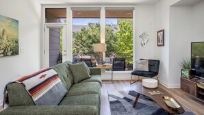 A modern living room with a sofa, a coffee table, a TV, and a view through large windows. - Beautiful 2-bedroom townhome downtown Raleigh  Walk to Everything (Raleigh)