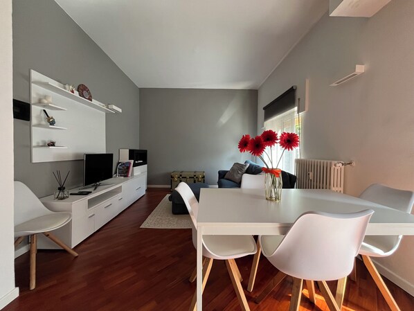 Uma sala de estar moderna com uma mesa de jantar, cadeiras brancas, um sofá, uma TV e um vaso com flores vermelhas.