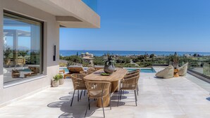 A modern outdoor patio with a wooden table and chairs, a potted plant, and a view of the sea. - Olea Hill Villas - Luxury Villa Lamina, by Estia (Vathianos Kampos)