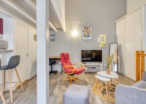 A modern living room with a flat-screen TV, a red chair, a grey sofa, and a small round table. - HEVA – Studio cosy en plein cœur de Rouen (Rouen)