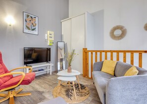 A modern living room with a TV, red chair, grey sofa, wooden headboard, and a small table with a vase. - HEVA – Studio cosy en plein cœur de Rouen (Rouen)