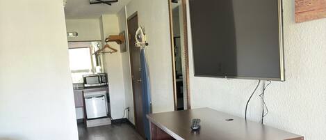 A compact hotel room with a bed, a desk with drawers, a television, and a kitchenette in the background.