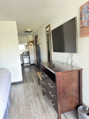 Habitación de hotel compacta con cama, escritorio con cajones, televisor y una kitchenette al fondo.